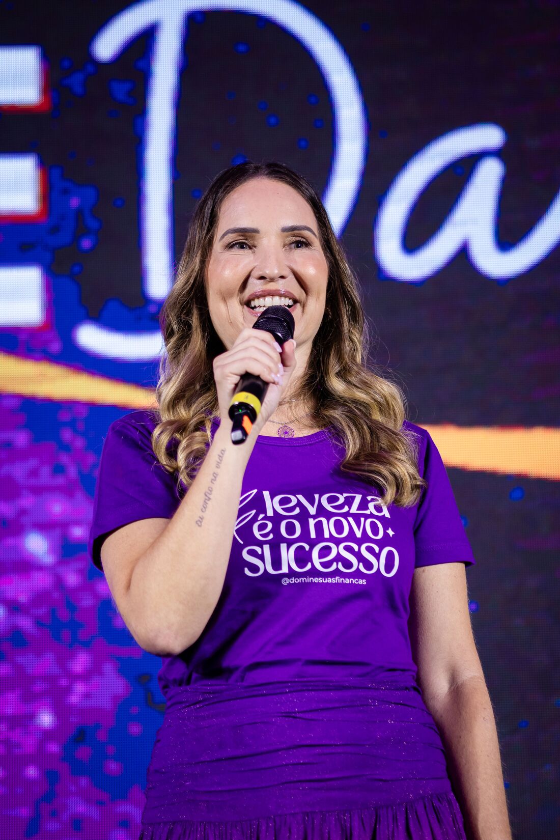 Luciana Fiaux falando ao microfone em um palco iluminado de roxo